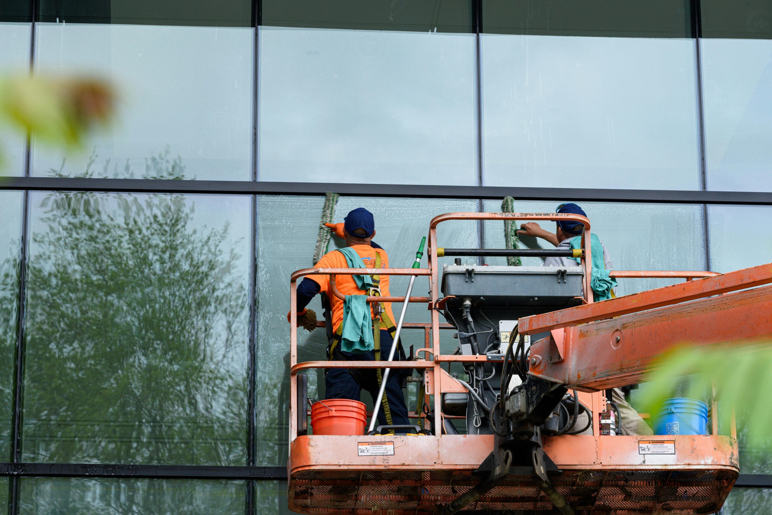 Two workers cleaning windows on a glass building facade using a lift platform, enhancing building aesthetics.