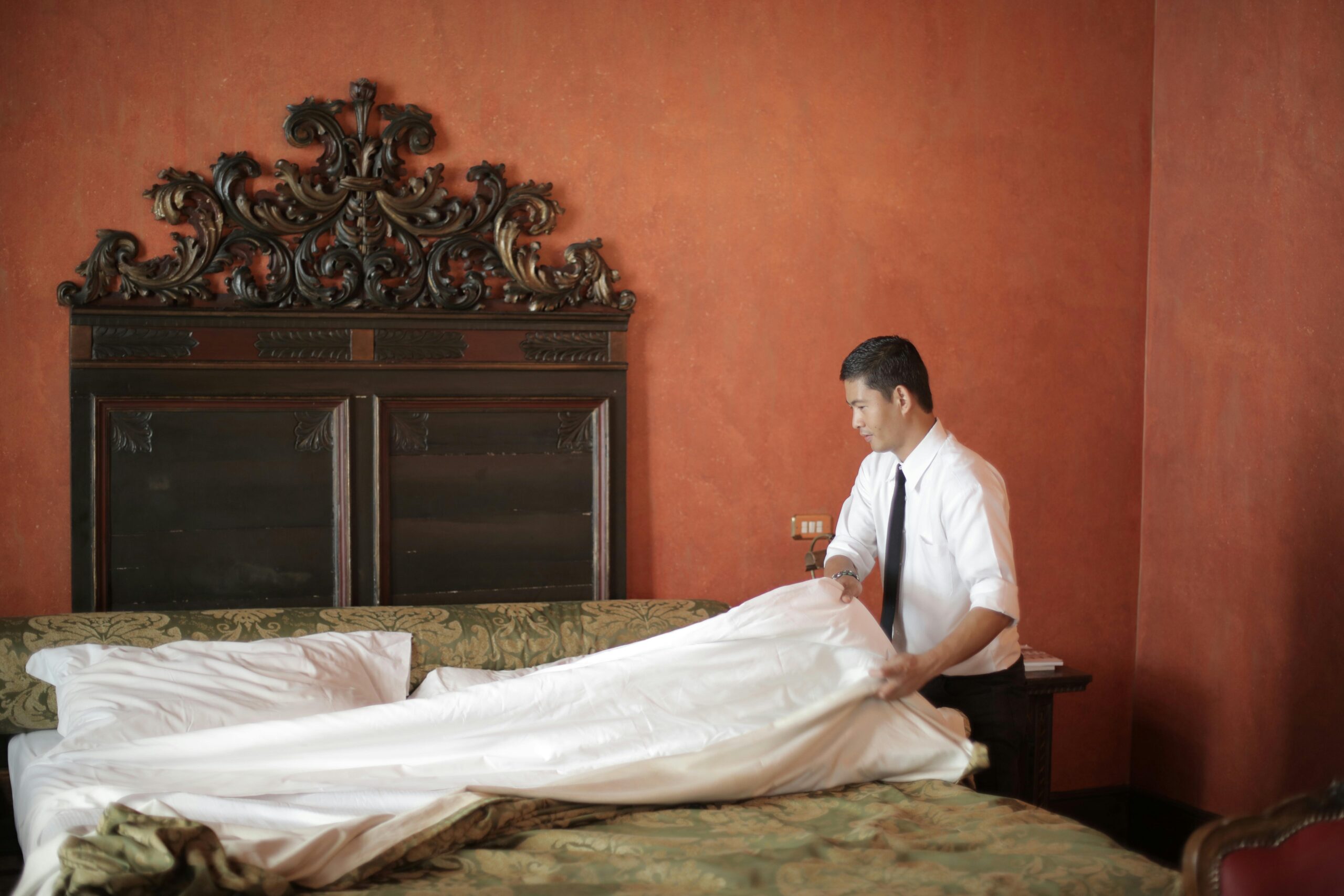 A hotel staff member in formal attire making a bed in a luxurious bedroom setting.