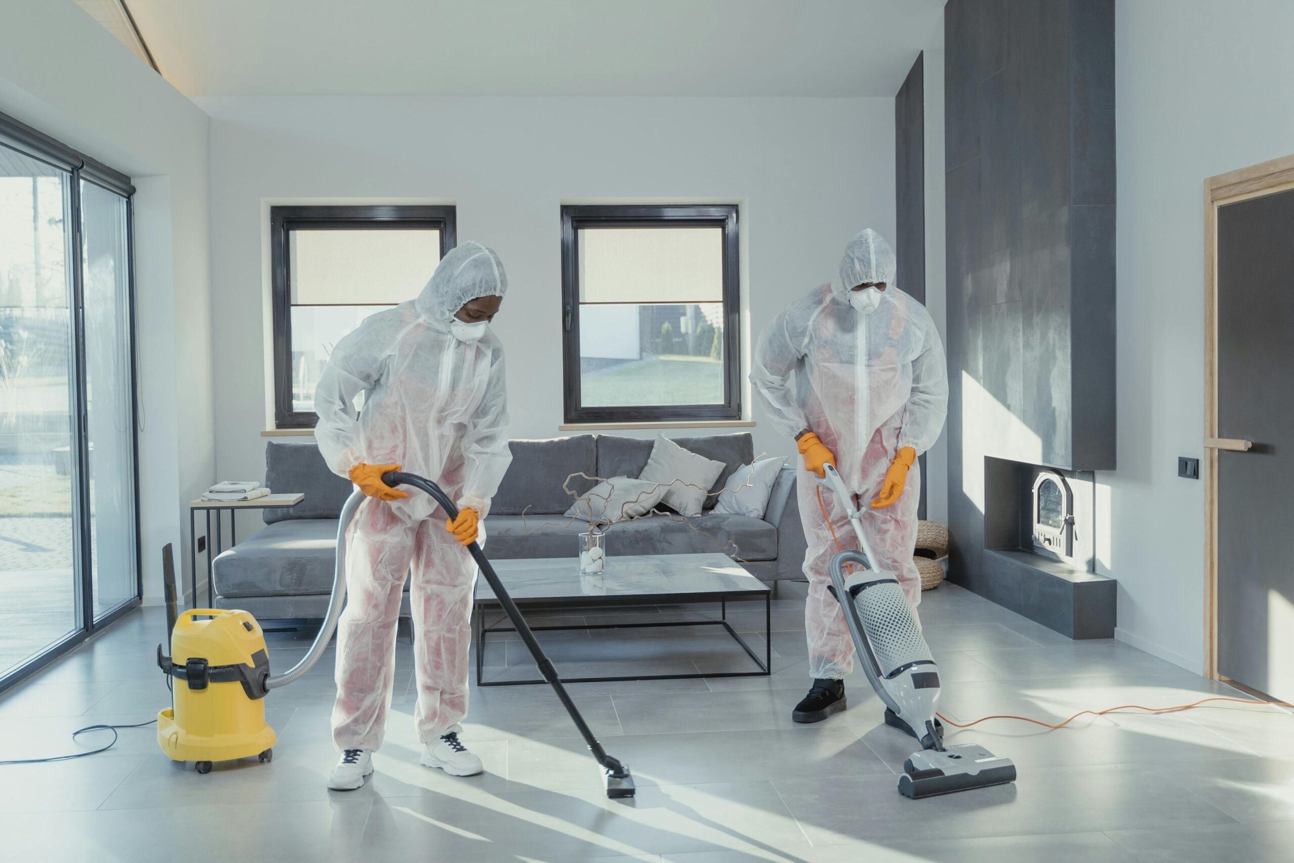 Two workers in protective gear vacuum and disinfect a modern living room.